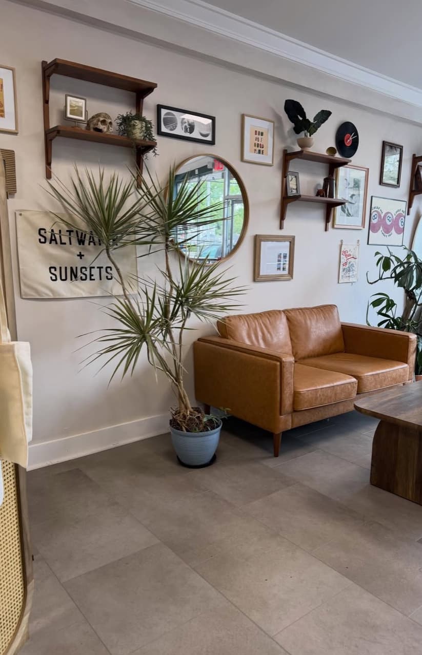 A gallery wall with a round mirror, framed art, and a leather couch, with a Saltwater + Sunsets banner