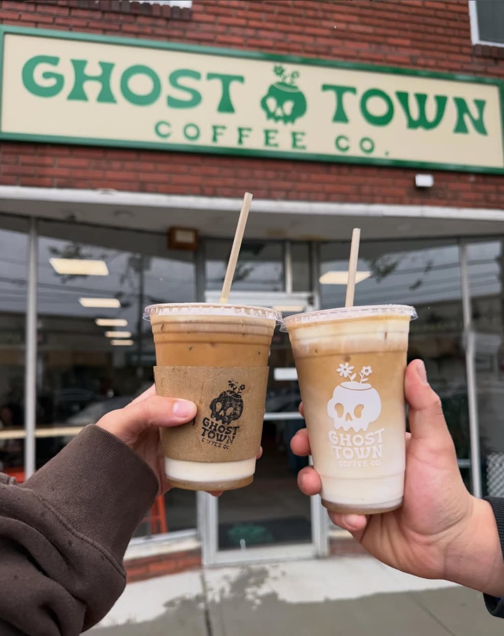 Two iced coffees held up in front of the Ghost Town Coffee Co. storefront sign