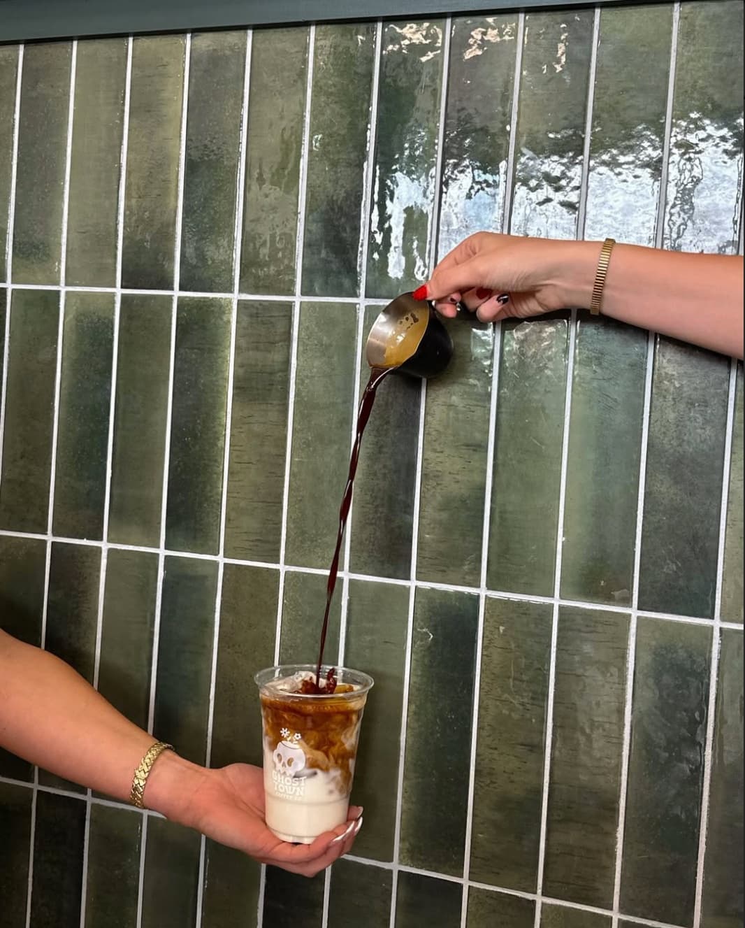 An iced coffee in a Ghost Town Coffee Co. cup being finished with a pour, against a green tiled wall